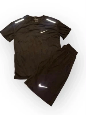 Svart träningsset från Nike - Säljer ett svart träningsset från Nike med t-shirt och shorts. Båda plaggen har vita Nike-loggor och reflekterande detaljer på axlar och ben. Perfekt för gymmet eller löpning. Materialet är lätt och andas, vilket gör det bekvämt under träning.