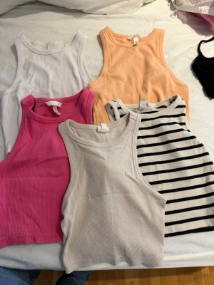 Fem ribbade linnetoppar i olika färger - Säljer ett paket med fem ribbade linnetoppar i olika färger: vit, beige, rosa, orange och en vit med svarta ränder. Alla är ärmlösa och har en croppad passform, perfekta för sommaren. Snygga att matcha med jeans eller kjol! alla är i storleken S förutom den Beige som är i Xs. 