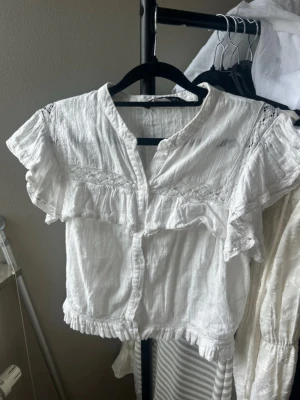 Vit blus med volanger från Zara - Söt vit blus från Zara med volangdetaljer och spets längs axlar och bröst. Blusen har knappar framtill, korta ärmar med volanger och är tillverkad i ett lätt, luftigt tyg som känns som bomull. Perfekt för en trendig och avslappnad look.