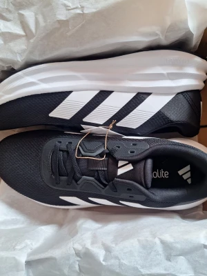 Svarta sneakers med vita ränder - Snygga svarta sneakers från Adidas med klassiska vita ränder på sidan och vit sula. Ovandelen är i meshmaterial för bra andningsförmåga och komfort. Perfekta för en sportig och avslappnad stil. Modellen har snörning och lätt vadderad plös.
