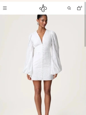Vit florence kort klänning med broderi från Adoore - Stilren vit kortklänning med v-ringning, broderade detaljer och långa puffärmar. Klänningen har knappar framtill och markerad midja som ger en feminin siluett. Perfekt för dig som vill ha en elegant och trendig look. Klänning från adoore vit florence 
