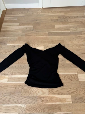 Svart offshoulder topp - Snygg svart omlottopp med offshoulder-design och långa ärmar. Toppen har en figurnära passform och är tillverkad i ett mjukt, stretchigt material som ger en bekväm känsla. Perfekt för dig som vill ha en stilren och trendig look. Djup i ryggen som gör att den passar både till fest ich vardags. Använd ca 1gg på fest❣️