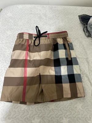 Rutiga badbyxor med snörning - Snygga badbyxor med klassiskt rutigt mönster i beige, brunt, svart, vitt och rött. De har elastisk midja med röd kant och svart snörning samt ficka med dragkedja på sidan. Perfekta för stranden eller poolen.