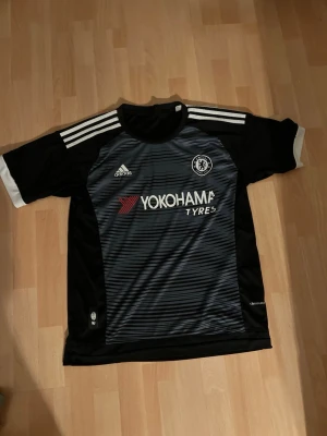 Chelsea FC svart matchtröja Adidas - Snygg svart Chelsea FC matchtröja från Adidas med vita detaljer och ränder på axlarna. Framsidan har Yokohama Tyres-logga och klubbmärke. Tillverkad i lätt och ventilerande material, perfekt för fotboll eller träning.