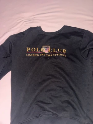 Svart långärmad topp från Vinson Polo Club - Svart långärmad topp från Vinson Polo Club med broderad logga och texten 'Legendary Traditions' i guld på bröstet. Rund halsringning med en diskret vinröd detalj på insidan av kragen. Perfekt för en stilren och sportig look.