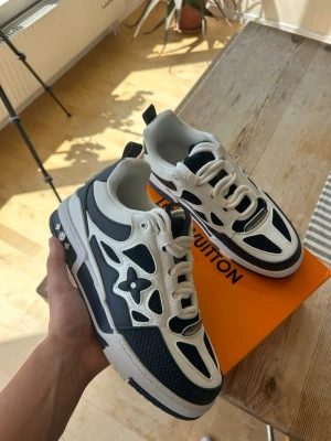 Louis Vuitton vita och marinblå sneakers - Snygga Louis Vuitton sneakers i vitt och marinblått med mesh och läderdetaljer. Skorna har en chunky sula, LV-logga på sidan och coola stjärndetaljer. Perfekta för dig som gillar streetwear och vill sticka ut med exklusiva detaljer.
