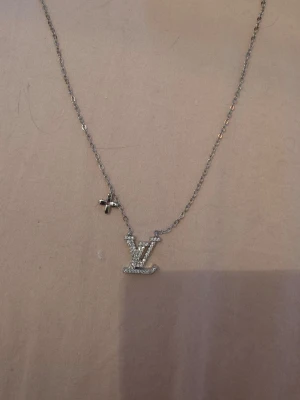 Louis Vuitton halsband  - Snyggt halsband i silverfärg med ett glittrande LV-hänge. Kedjan är tunn och elegant, och hänget är täckt av små stenar som ger extra bling. Perfekt för dig som gillar stilrena accessoarer med en lyxig touch.