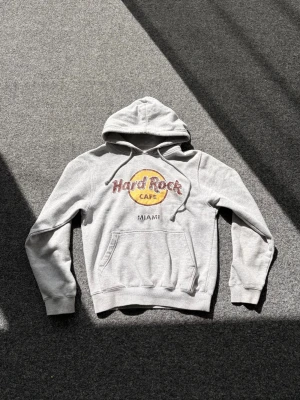 Grå Hard Rock Cafe hoodie Miami - Säljer en grå hoodie med Hard Rock Cafe Miami-tryck i vinrött och gult på bröstet. Klassisk känguruficka framtill och justerbar huva med dragsko. Perfekt för dig som gillar ikoniska tryck och avslappnad stil.