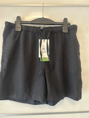 Svarta linneshorts med snörning - Snygga svarta shorts i linne med avslappnad passform och elastisk midja med snörning. Perfekta för varma dagar och ger en clean och enkel look. Säljer pga jag köpte i för liten storlek.