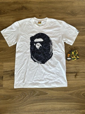 Vit BAPE t-shirt med stor logga - Vit t-shirt från BAPE med stor svart logga i form av ett apehuvud på framsidan och en mindre logga med texten '30th Anniversary' på ryggen. Klassisk passform och rund hals. Tillverkad i mjuk bomull och levereras med originaltaggar.