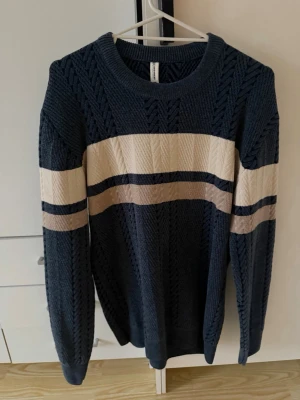 Mörkblå kabelstickad tröja med ränder - Snygg mörkblå kabelstickad tröja med breda beige och vita ränder över bröstet och armarna. Tröjan har rund halsringning och långa ärmar. Perfekt för dig som gillar klassisk men trendig stil.
