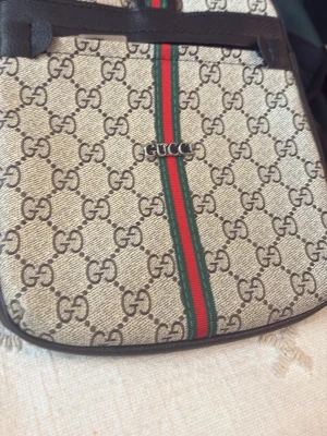  axelväska med GG-mönster - Snygg axelväska  med klassiskt GG-monogram i beige och brunt, samt den ikoniska gröna och röda randen framtill. Väskan har detaljer i mörkbrunt läder och justerbar axelrem. Baksidan är helsvart i slätt läder. Perfekt för dig som gillar lyxig och tidlös design. Har inte andvänd länge 
