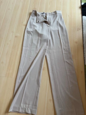 Beige vida kostymbyxor - Snygga beige kostymbyxor med hög midja och vida ben. Byxorna har en stilren design med knapp- och dragkedjeförslutning framtill. Perfekta för en clean och modern look. Materialet känns mjukt och följsamt.