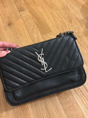 Svart YSL handväska i läder - Snygg svart handväska från YSL i quiltat läder med silverfärgad kedjerem och ikonisk YSL-logga framtill. Väskan har ett klassiskt rektangulärt snitt och magnetlås. Perfekt för dig som gillar tidlös design och vill ha en statementväska.