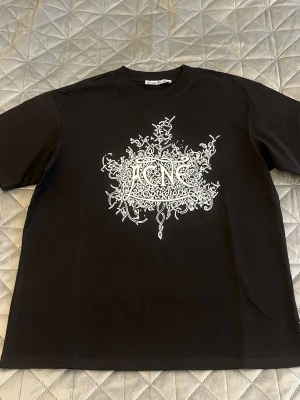 Svart Acne Studios t-shirt med tryck - Säljer en svart t-shirt från Acne Studios med ett stort vitt grafiskt tryck på bröstet. T-shirten har rund hals och korta ärmar. Perfekt för dig som gillar streetwear och vill ha något unikt i garderoben.