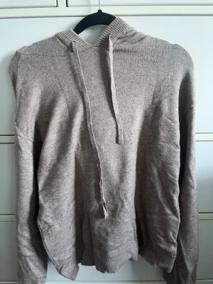 Beige stickad hoodie med huva - Mysig beige stickad hoodie med huva och dragsnören. Tröjan har långa ärmar och ribbade muddar vid ärmslut och nederkant. Perfekt för en avslappnad och chill stil. Materialet känns mjukt och bekvämt mot huden.