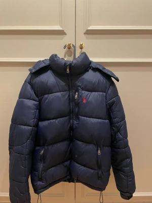 Mörkblå pufferjacka från Polo Ralph Lauren - Snygg mörkblå pufferjacka från Polo Ralph Lauren med röd logga på bröstet. Jackan har hög krage, avtagbar huva och flera dragkedjeförsedda fickor. Perfekt för kalla vinterdagar och riktigt skön att bära. Klassisk quiltad design och sportig look.