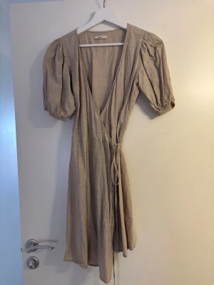 Beige linneklänning med puffärm - Snygg beige omlottklänning i linne-look med puffiga korta ärmar och knytband i midjan. Djup V-ringning och avslappnad passform ger en trendig och luftig känsla. Perfekt för dig som gillar stilrena och enkla plagg med en twist.