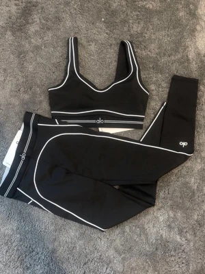 ALO träningsset  - Snyggt svart träningsset från Alo med vita kontrastsömmar. Setet består av en sport-bh med breda axelband och ett par tights med hög midja. Materialet är stretchigt och följsamt, perfekt för gymmet eller yoga. 