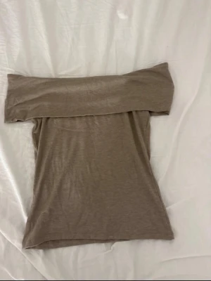 Beige offshoulder topp från Kappahl - Säljer en beige offshoulder topp från Kappahl i mjukt och stretchigt material. Toppen har en bred kant upptill som ger en snygg axelbandslös look och passar perfekt till både jeans och kjol. Enkel och stilren design som är lätt att matcha.