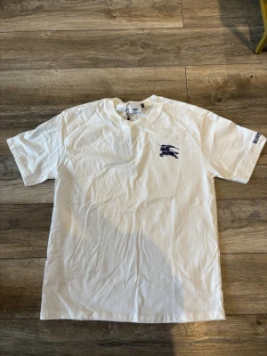 Vit Burberry t-shirt med logga - Säljer en vit t-shirt från Burberry med klassisk logga på bröstet och Burberry-tryck på ärmen. T-shirten har rund hals och korta ärmar. Perfekt basplagg med exklusiv känsla och stilren design.
