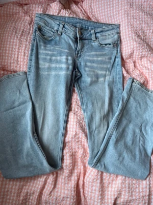 Ljusblå raka jeans med tryckt patch - Säljer ett par ljusblå jeans med raka ben och klassisk femficksmodell. Jeansen har slitningar framtill och en stor vit patch med tryck bak i midjan. Materialet är denim i bomull och passformen är relaxed. Perfekta för en avslappnad streetstil.