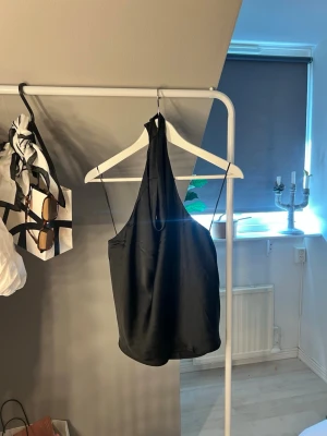 Svart halterneck topp i satin - Snygg svart linnetopp i satin med halterneck och tunna axelband. Toppen har en draperad framsida och är croppad, vilket ger en trendig och stilren look. Perfekt att matcha med jeans eller kjol för en cool outfit.