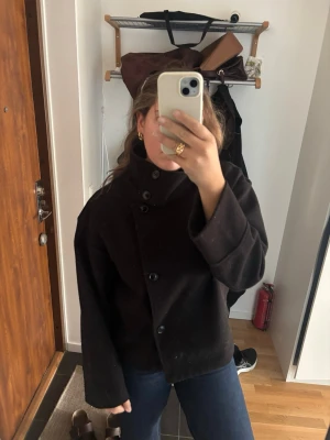 Svart kappa med hög krage - Stilren svart kappa i ullmix med hög krage och asymmetrisk knäppning framtill. Modellen är boxig med vida ärmar och knappar längs ena sidan. Perfekt för dig som gillar minimalistisk och modern stil.