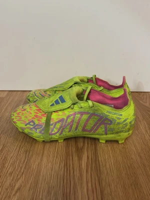 Adidas Predator neon fotbollsskor - Säljer ett par Adidas Predator fotbollsskor i neonfärg med rosa och blå detaljer. Skorna har en låg profil, snörning och täckt plös. Insidan är rosa och yttermaterialet är syntetiskt med mönstrad textur för bättre bollkontroll. Perfekta för dig som vill sticka ut på planen.