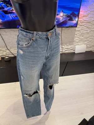Ljusa slitna raka jeans - Säljer ett par ljusblå jeans med raka ben och slitningar vid knäna och låret. Jeansen har klassisk femficksmodell, hög midja och är tillverkade i denim. Perfekta för en avslappnad och trendig look.