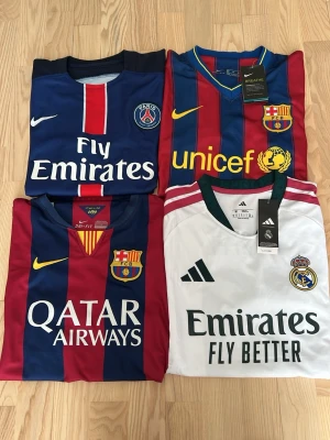 Fotbollströjor PSG, Barcelona, Real Madrid - Fyra fotbollströjor: PSG i blått/rött med Ibrahimović 10, två Barcelona-tröjor i rödblått med Neymar Jr 11 och klubbmärke, samt en vit Real Madrid-tröja med gröna detaljer och Mbappé 10. Alla har tryckta sponsorer och klubbmärken. Perfekt för fotbollsfans!
