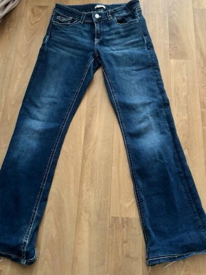 Mörkblå bootcut jeans med fickdetaljer - Dessa jeans fixa jag för personer som är runt 148-155 cm långa. Snygga mörkblå bootcut jeans med slitningar och kontrastsömmar. Jeansen har framfickor och bakfickor med lock och knappdetaljer. Klassisk femficksmodell i mjukt denimtyg, perfekta för en avslappnad och trendig look. 