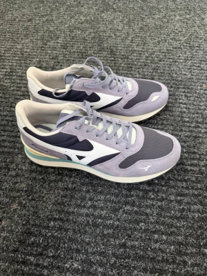 Lila och vita sneakers från Mizuno - Snygga sneakers i lila, vitt och mörkblått från Mizuno. Skorna har en klassisk låg siluett, mesh- och mockadetaljer samt en vit sula med pastellfärgade inslag. Perfekta för dig som gillar sportig och stilren design.