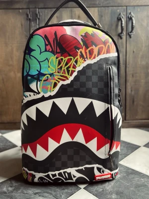 Sprayground ryggsäck Limited Edition  - Säljer en unik ryggsäck från Sprayground med coolt graffiti- och hajmotiv. Väskan har svart och grått rutmönster, färgglada detaljer och stora vita tänder över framsidan. Tillverkad i slitstarkt material med flera fack och dragkedjor. Mått- 45x15x29cm. Endast använd en gång. Som ny, 