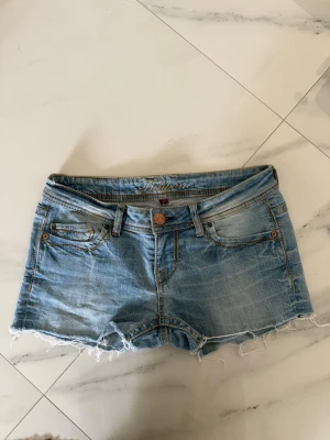 Ljusblå jeansshorts från Fishbone - Säljer ett par ljusblå jeansshorts från Fishbone med fransig kant och klassisk femficksmodell. Shortsen har bälteshällor, knappgylf och slitna detaljer för en avslappnad look.