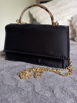 Svart kuvertväska med gulddetaljer - Stilren svart kuvertväska i satinliknande material med elegant guldfärgat handtag och avtagbar kedja. Perfekt för att lyfta din outfit med en lyxig touch. Väskan har en klassisk rektangulär form och stängs med lock.