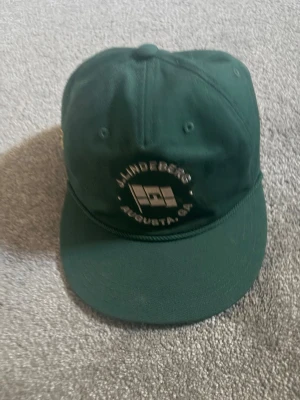 Mörkgrön keps från J.Lindeberg - Snygg mörkgrön keps från J.Lindeberg med broderad logga och texten 'AUGUSTA GA' framtill. Klassisk form med böjd skärm och justerbar baksida. Tillverkad i 100% bomull för en skön och luftig känsla.