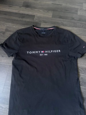 Svart T-shirt från Tommy Hilfiger - Snygg svart T-shirt från Tommy Hilfiger med klassisk logga och texten 'EST. 1985' på bröstet. Tillverkad i ekologisk bomull och har en normal passform med korta ärmar. Perfekt till jeans eller shorts för en clean look.