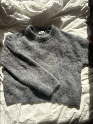 Grå fluffig stickad tröja - Mysig grå stickad tröja med fluffig yta och rund hals. Tröjan har en avslappnad passform och långa ärmar, perfekt för kyliga dagar. Materialet ser ut att vara en mjuk ull- eller mohairblandning.