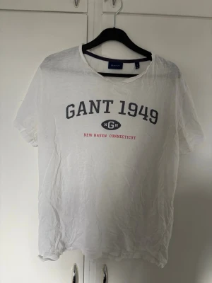 Vit GANT t-shirt - Vit t-shirt från GANT med trycket 'GANT 1949' och 'New Haven Connecticut' på bröstet. Klassisk rund halsringning och korta ärmar. Tillverkad i mjukt bomullsmaterial, perfekt för en avslappnad stil. Urtvättad och sliten men en väldigt fin t shirt