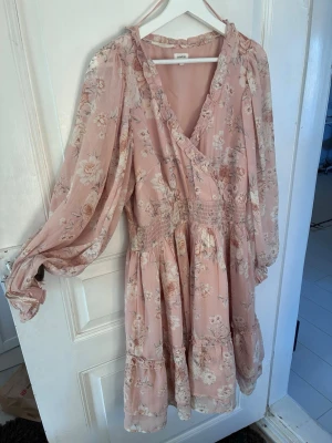 Blommig rosa omlottklänning Newbie M - Supersöt rosa omlottklänning från Newbie i storlek M. Klänningen har blommigt mönster, volangdetaljer, långärmade puffärmar och smock i midjan. Perfekt för vår och sommar med sitt lätta, luftiga tyg och romantiska look.