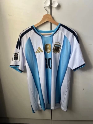 Argentina Messi matchtröja - Säljer en klassisk Argentina matchtröja med Messi 10 på ryggen. Tröjan har blå och vita vertikala ränder, svarta detaljer och guldiga loggor på bröstet. Tillverkad i lätt och ventilerande material, perfekt för fotboll eller streetwear.