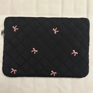 Svart quiltad datorväska med rosetter från SHEIN - Svart quiltad datorväska med dragkedja och små rosa rosetter broderade på framsidan från SHEIN. Väskan har en mjuk vadderad känsla och ett rutigt mönster i tyget. Måtten på datorväskan: ca 24 cm lång och 35 cm bred. OBSERVERA att det kan förekomma små fläckar på väskan vid nära anblick. 