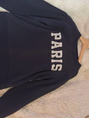 Mörkblå sweatshirt med PARIS-tryck - Mörkblå sweatshirt med stora, vita bokstäver som stavar PARIS på bröstet och texten 'Que des artistes' under. Tröjan har rund halsringning och långa ärmar. Perfekt för en avslappnad och trendig look.