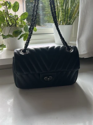 Svart quiltad axelremsväska - Snygg svart handväska i quiltat mönster med kedjerem och silverfärgade detaljer. Väskan har ett klassiskt lock med vridlås och flera fack inuti för bra organisering. Perfekt storlek för det viktigaste och ger en stilren look.