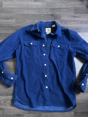 Blå manchesterskjorta från Levi's - Säljer en blå manchesterskjorta från Levi's i modellen Jackson Worker. Skjortan har avslappnad passform, två bröstfickor med lock och vita knappar framtill. 