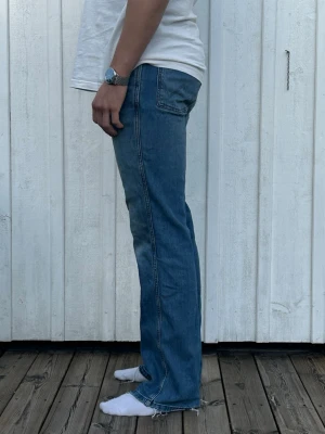 Wrangler WRANCHER - Säljer ett par blå bootcut jeans med lätt slitna detaljer vid benslut ifrån Wrangler. Dom är relativt små i storleken