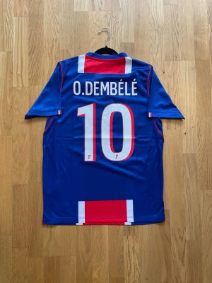 O. Dembélé #10 - S - Säljer en officiell Paris Saint-Germain matchtröja från Nike med O. Dembélé och nummer 10 på ryggen. Tröjan är blå med röda och vita detaljer, PSG-logga och Qatar Airways sponsortryck på bröstet. Korta ärmar och sportig passform, perfekt för fotbollsfans.