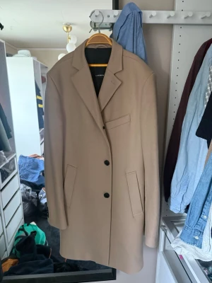 Beige ullkappa från J.Lindeberg - Stilren beige kappa från J.Lindeberg i ullblandning. Klassisk rak modell med tre knappar framtill, två sidofickor och en bröstficka. Perfekt för dig som vill ha en tidlös och snygg look till hösten eller vintern.