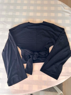 Svart topp med knyt - Snygg svart topp med långa ärmar och knyt i ryggen. Toppen har en enkel, stilren design och är tillverkad i mjukt material som känns skönt mot huden. Perfekt för att lyfta vilken outfit som helst.